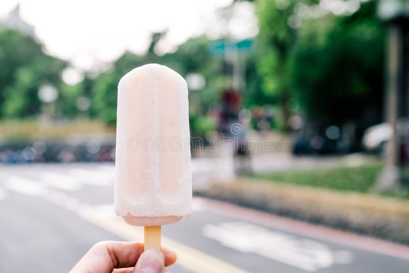 Hot summer pink popsicle stick with street background. Hand is holding the ice cream. Hot summer pink popsicle stick with street background. Hand is holding the ice cream.