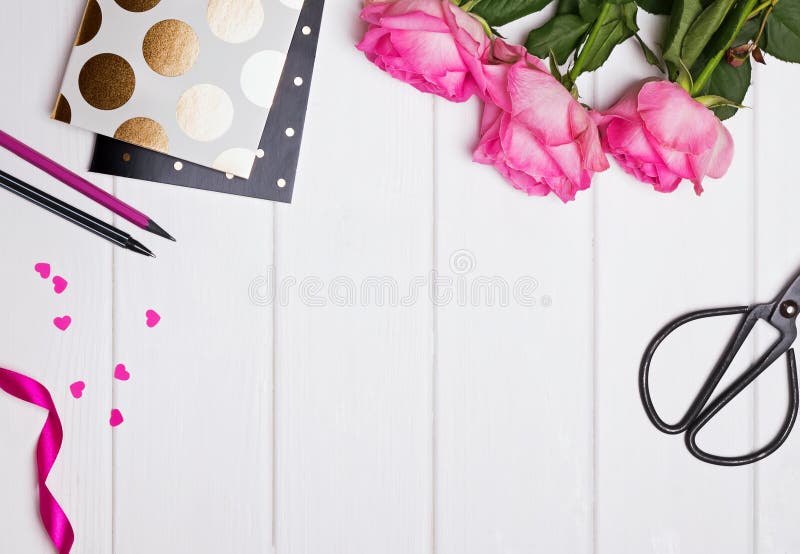 Styled desk. Pink roses, notepads with gold polka dot, paper hearts and other objects on the white wooden table, top view.