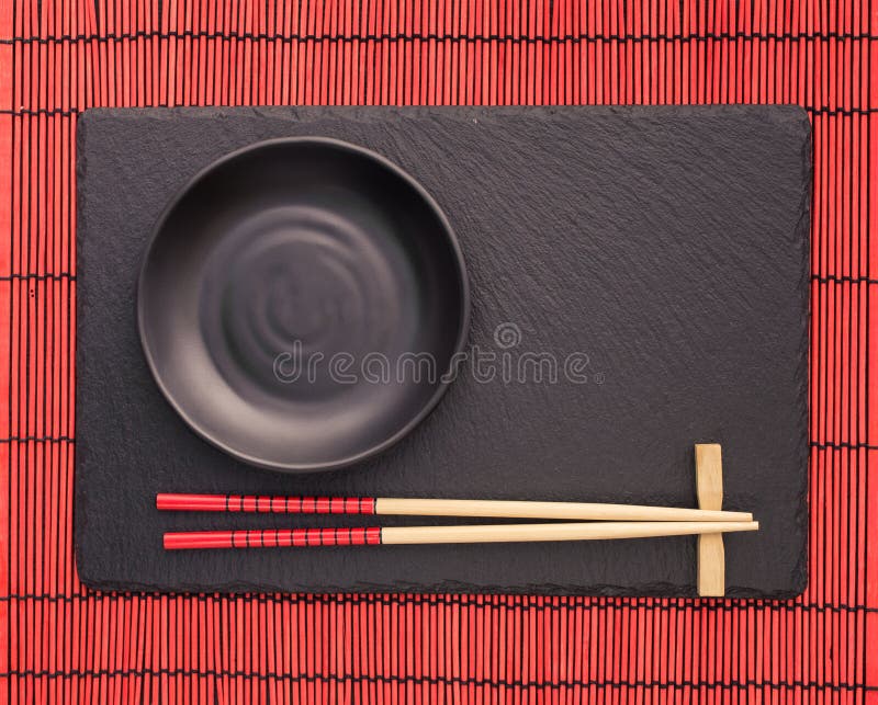 Chopsticks and black plate on slate coaster on red bamboo scroll mat. Japanese style food background. Top view with copy space. Chopsticks and black plate on slate coaster on red bamboo scroll mat. Japanese style food background. Top view with copy space