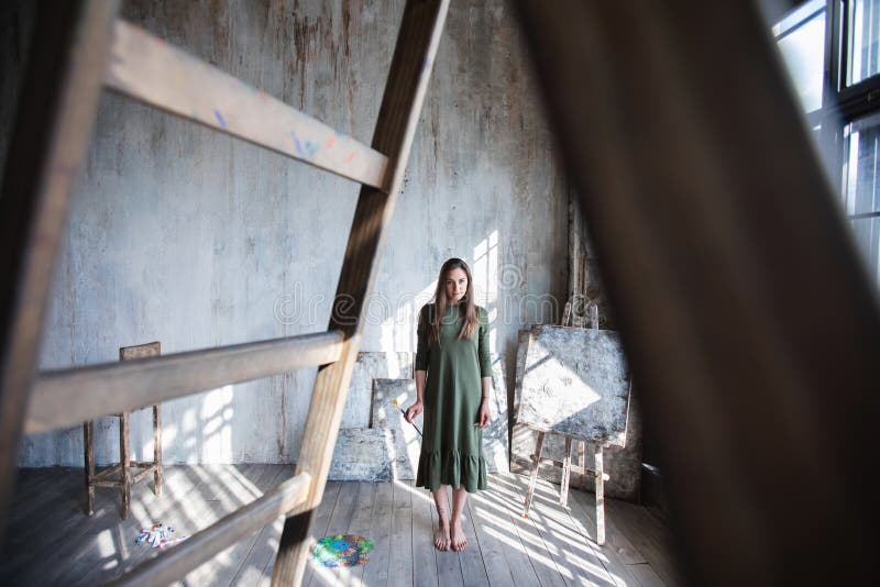 Pintor de pelo largo joven de la mujer en un vestido verde con un cepillo y una paleta en un estudio del arte fotografía de archivo Pintor de pelo largo joven de la mujer en un vestido verde con un cepillo y una paleta en un estudio del arte fotografía de archivo