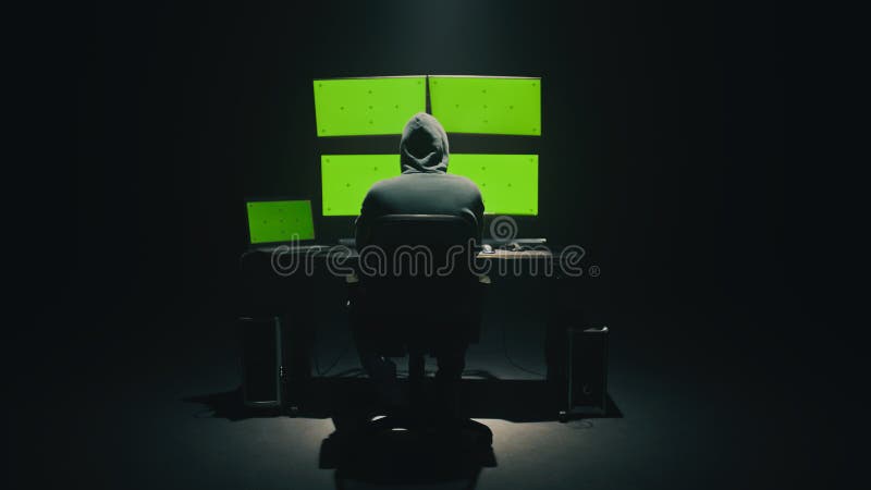 Concept shot of anonymous man in hoodie writing code to hack database while sitting at desk with computer monitors, with green screens chromakey in dim room of hacker base. Concept shot of anonymous man in hoodie writing code to hack database while sitting at desk with computer monitors, with green screens chromakey in dim room of hacker base