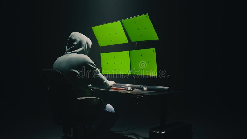 Concept shot of anonymous man in hoodie writing code to hack database while sitting at desk with computer monitors, with green screens chromakey in dim room of hacker base. Concept shot of anonymous man in hoodie writing code to hack database while sitting at desk with computer monitors, with green screens chromakey in dim room of hacker base