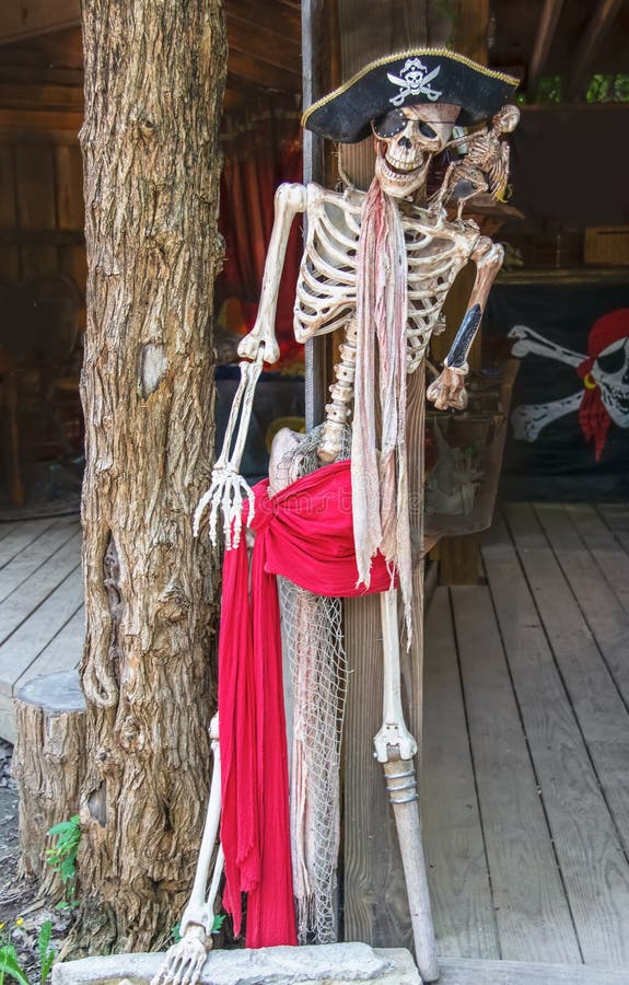 Piratkopiera skelettet med pinnebenet, och ögonlappen och hatten och den döda papegojan på skuldran propped på pelare för trädsta royaltyfri fotografi Piratkopiera skelettet med pinnebenet, och ögonlappen och hatten och den döda papegojan på skuldran propped på pelare för trädsta royaltyfri fotografi