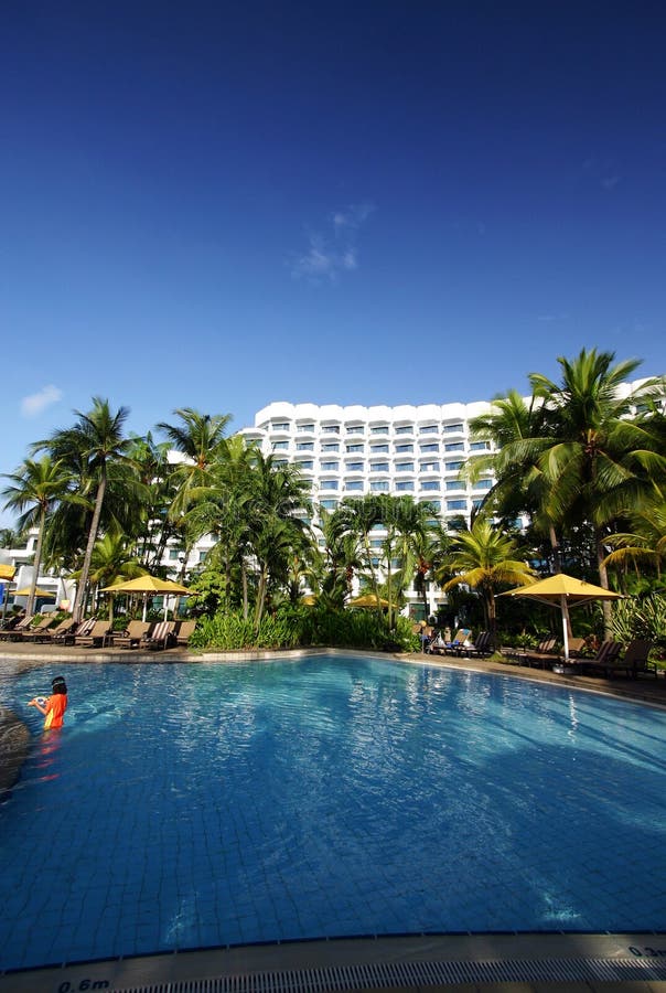 An image showing the beautiful architecture of a modern tropical beach resort, with the landscaped swimming pool in the foreground and stylish white resort building in background. Taken under an azure deep blue tropical sky. Palm trees in the lush landscaping and green tropical plants add to the exotic tropics mood of the garden grounds. With bright yellow sunshades beside the pool and rows of relaxing sun bed loungers. Child in orange swimsuit playing in pool. Vertical color format. Taken at a five star luxury resort hotel in Sentosa Island, Singapore, southeast asia. Landscaped garden with palms. Taken with little girl child playing in water. Fun vacation for kids. An image showing the beautiful architecture of a modern tropical beach resort, with the landscaped swimming pool in the foreground and stylish white resort building in background. Taken under an azure deep blue tropical sky. Palm trees in the lush landscaping and green tropical plants add to the exotic tropics mood of the garden grounds. With bright yellow sunshades beside the pool and rows of relaxing sun bed loungers. Child in orange swimsuit playing in pool. Vertical color format. Taken at a five star luxury resort hotel in Sentosa Island, Singapore, southeast asia. Landscaped garden with palms. Taken with little girl child playing in water. Fun vacation for kids.