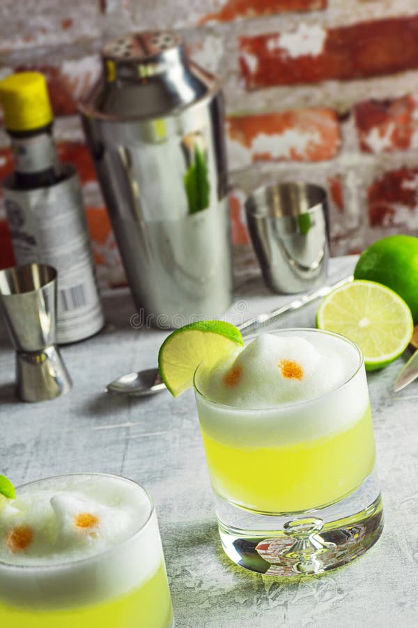 Two rocks glasses filled with pisco sours and garnished with lime wedges on a bar top. This cocktail is made from Peruvian pisco liquor, lime juice, simple syrup, and egg white shaken together and topped with a couple dashes of bitters. The tools and ingredients used to make the drinks are in the background. Two rocks glasses filled with pisco sours and garnished with lime wedges on a bar top. This cocktail is made from Peruvian pisco liquor, lime juice, simple syrup, and egg white shaken together and topped with a couple dashes of bitters. The tools and ingredients used to make the drinks are in the background.