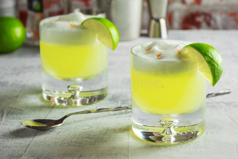 Two rocks glasses filled with pisco sours and garnished with lime wedges on a bar top. This cocktail is made from Peruvian pisco liquor, lime juice, simple syrup, and egg white shaken together and topped with a couple dashes of bitters. The tools and ingredients used to make the drinks are in the background. Two rocks glasses filled with pisco sours and garnished with lime wedges on a bar top. This cocktail is made from Peruvian pisco liquor, lime juice, simple syrup, and egg white shaken together and topped with a couple dashes of bitters. The tools and ingredients used to make the drinks are in the background.