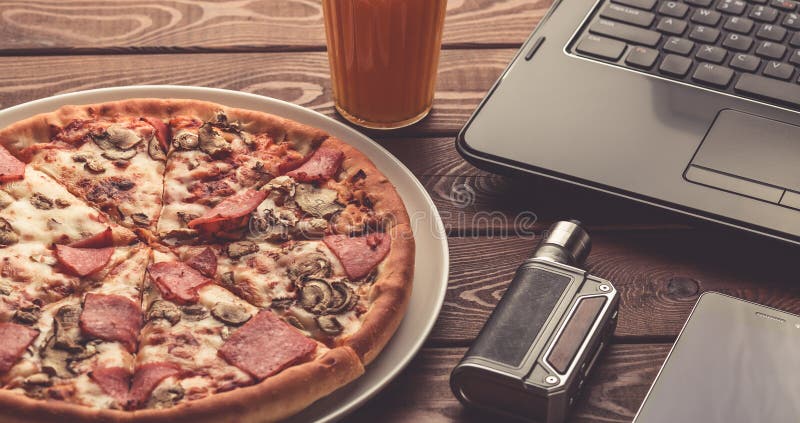 Pizza su un piatto, un computer portatile nero, una sigaretta o un vape elettronico, un telefono cellulare e un vetro del succo d fotografia stock libera da diritti Pizza su un piatto, un computer portatile nero, una sigaretta o un vape elettronico, un telefono cellulare e un vetro del succo d fotografia stock libera da diritti