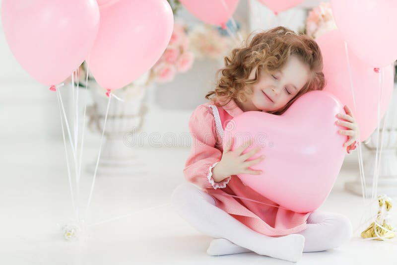 Valentine`s Day, postcard. Small girl with blond curly hair poses in a white studio surrounded by pink balloons. A beautiful preschool girl in a white studio with heart-shaped pink balloons. Valentine`s Day, love, birthday. Valentine`s Day, postcard. Small girl with blond curly hair poses in a white studio surrounded by pink balloons. A beautiful preschool girl in a white studio with heart-shaped pink balloons. Valentine`s Day, love, birthday.