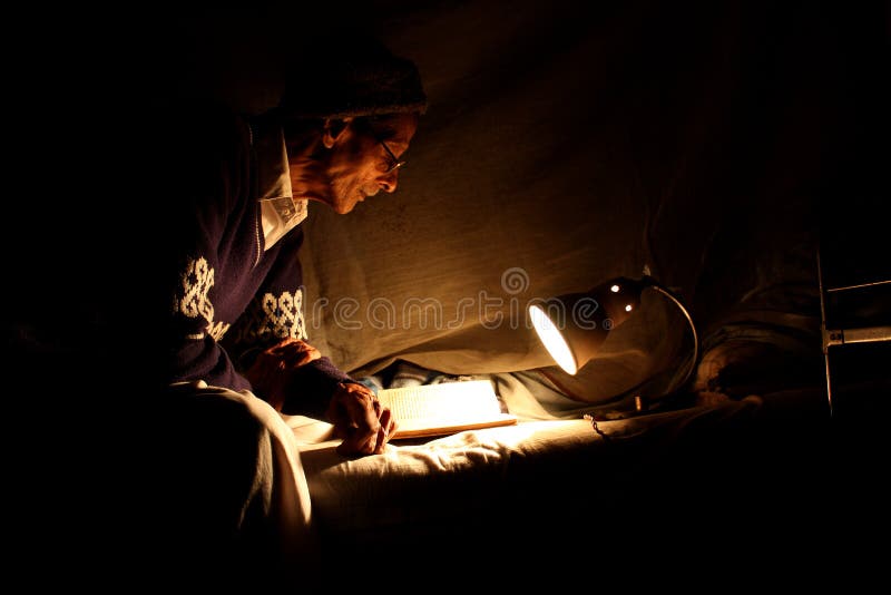 An old Indian man doing his research for thesis in the light of his retro table lamp on his bed. An old Indian man doing his research for thesis in the light of his retro table lamp on his bed