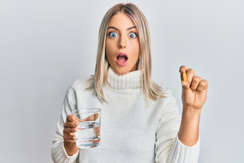 Beautiful blonde woman holding pill and glass of water afraid and shocked with surprise and amazed expression, fear and excited face. Beautiful blonde woman holding pill and glass of water afraid and shocked with surprise and amazed expression, fear and excited face