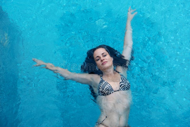 Piękna dojrzała kobieta w jej najlepszy wieku z ciemnym kędzierzawym włosy w bikini pływa z rękami podnosił weightless elegancki zdjęcia stock Piękna dojrzała kobieta w jej najlepszy wieku z ciemnym kędzierzawym włosy w bikini pływa z rękami podnosił weightless elegancki zdjęcia stock