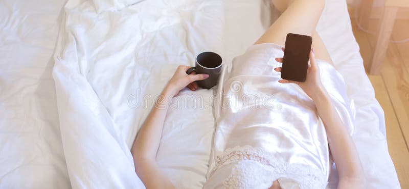 Beautiful girl is lying on white bed linen in grey loft minimalist interior. Young woman in silk robe is drinking coffee and reading news on internet. Social networks morning. Soft focus. Beautiful girl is lying on white bed linen in grey loft minimalist interior. Young woman in silk robe is drinking coffee and reading news on internet. Social networks morning. Soft focus.