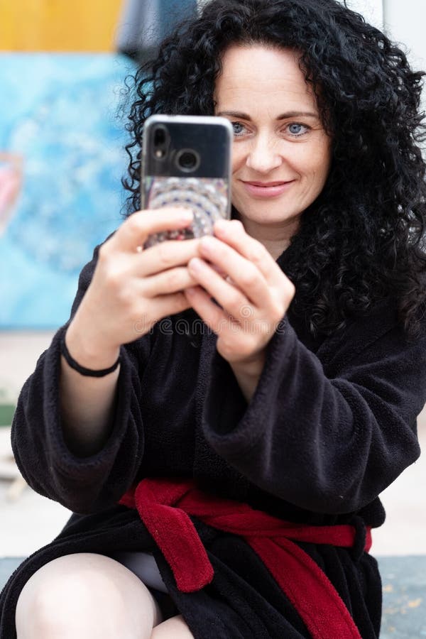 Piękna kobieta fotografuje ono uśmiecha się z smartphone z ciemnym kędzierzawym włosy w bathrobe zdjęcie royalty free Piękna kobieta fotografuje ono uśmiecha się z smartphone z ciemnym kędzierzawym włosy w bathrobe zdjęcie royalty free