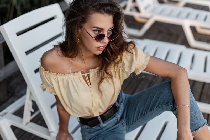 Piękna kobieta hipster z modnymi okularami słonecznymi w żółtej letniej bluzce z klasycznymi niebieskimi dżinsami siedzącymi i spo obraz stock