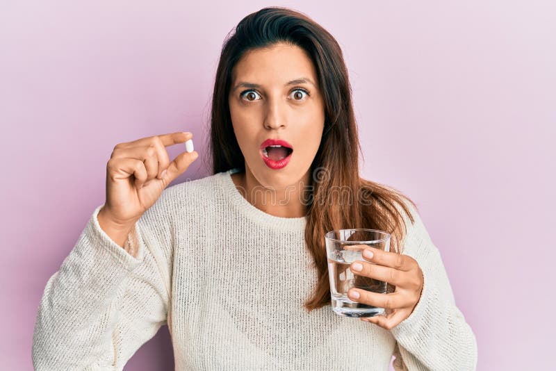 Beautiful hispanic woman holding pill and glass of water afraid and shocked with surprise and amazed expression, fear and excited face. Beautiful hispanic woman holding pill and glass of water afraid and shocked with surprise and amazed expression, fear and excited face
