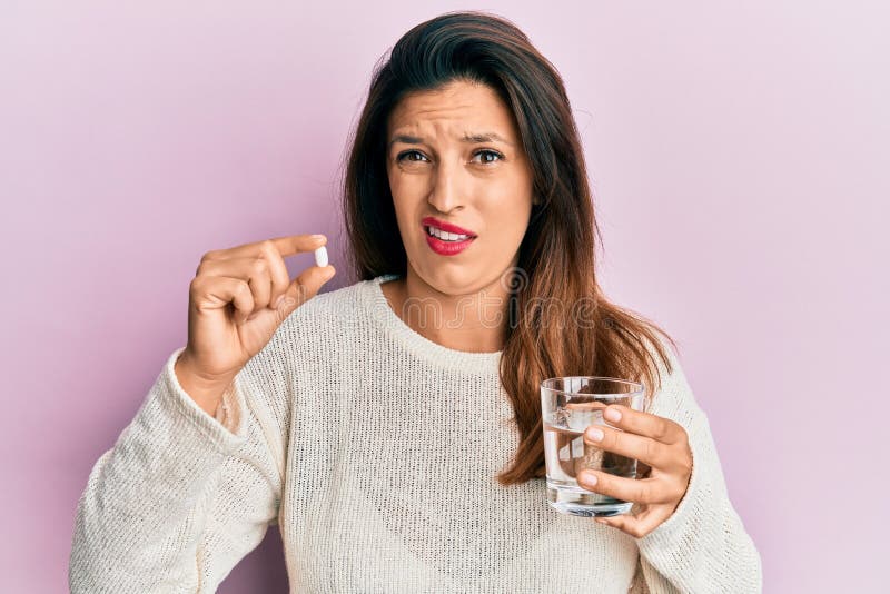 Beautiful hispanic woman holding pill and glass of water in shock face, looking skeptical and sarcastic, surprised with open mouth. Beautiful hispanic woman holding pill and glass of water in shock face, looking skeptical and sarcastic, surprised with open mouth