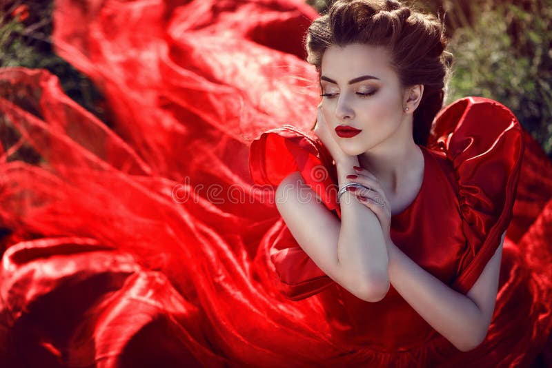 Beautiful young lady with perfect make up and plaited hairstyle wearing luxurious silk red ballroom dress sitting in the poppy field. The wind in her hair. The tail of her dress flying behind. Beautiful young lady with perfect make up and plaited hairstyle wearing luxurious silk red ballroom dress sitting in the poppy field. The wind in her hair. The tail of her dress flying behind