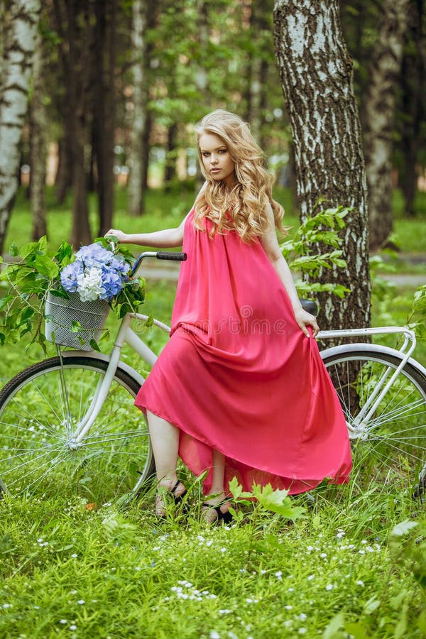 Piękna młoda dziewczyna w lato sukni przy zmierzchem Mody fotografia w lasowym modelu na bicyklu z kwiatu bukietem w menchii l, fotografia stock Piękna młoda dziewczyna w lato sukni przy zmierzchem Mody fotografia w lasowym modelu na bicyklu z kwiatu bukietem w menchii l, fotografia stock