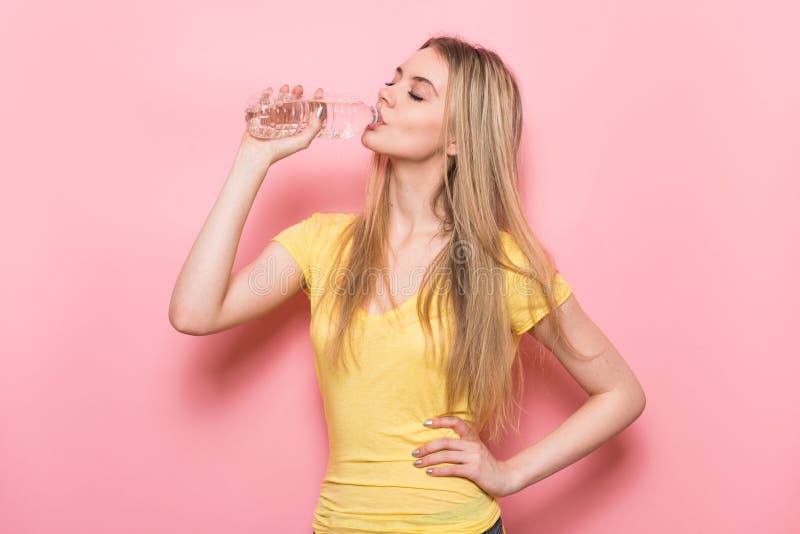 Piękna młoda sprawności fizycznej kobieta pije świeżą wodę od plastikowej butelki stoi blisko menchii ściany z perfect ciało kszt fotografia stock Piękna młoda sprawności fizycznej kobieta pije świeżą wodę od plastikowej butelki stoi blisko menchii ściany z perfect ciało kszt fotografia stock