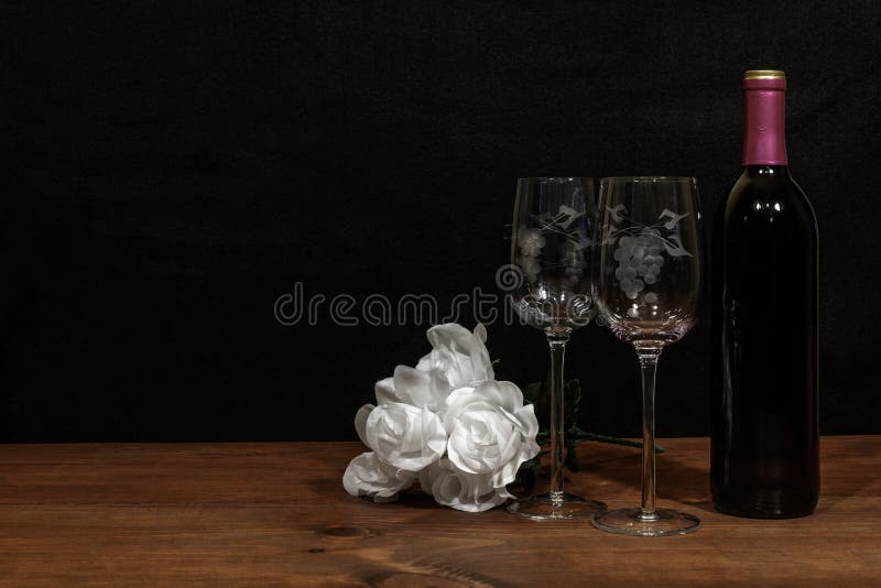 Beautiful etched wine glasses and bottle of red wine and white roses on wooden table and dark background. Valentines, Mothers Day, Easter, Christmas, Wedding Concepts. Beautiful etched wine glasses and bottle of red wine and white roses on wooden table and dark background. Valentines, Mothers Day, Easter, Christmas, Wedding Concepts