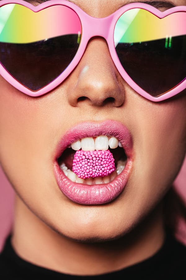 Closeup portrait of young woman wearing heart shaped sunglasses holding candy in mouth. Beautiful pink lips with a piece of sweet candy. Closeup portrait of young woman wearing heart shaped sunglasses holding candy in mouth. Beautiful pink lips with a piece of sweet candy.