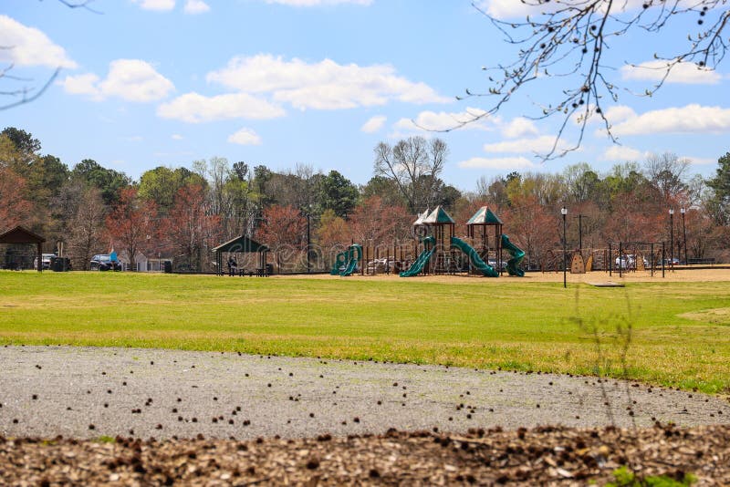 A gorgeous spring landscape in the park with a playground with a green and brown jungle gym, brown wooden pergolas, autumn colored trees, lush green trees, blue sky and clouds at Swift Cantrell Park in Kennesaw Georgia USA. A gorgeous spring landscape in the park with a playground with a green and brown jungle gym, brown wooden pergolas, autumn colored trees, lush green trees, blue sky and clouds at Swift Cantrell Park in Kennesaw Georgia USA