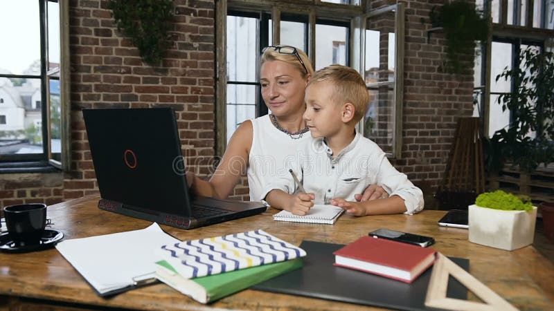 Piękny w średnim wieku bizneswoman pracuje na laptopie gdy jej mały uroczy wnuk coś napisze w notatniku przy zbiory wideo