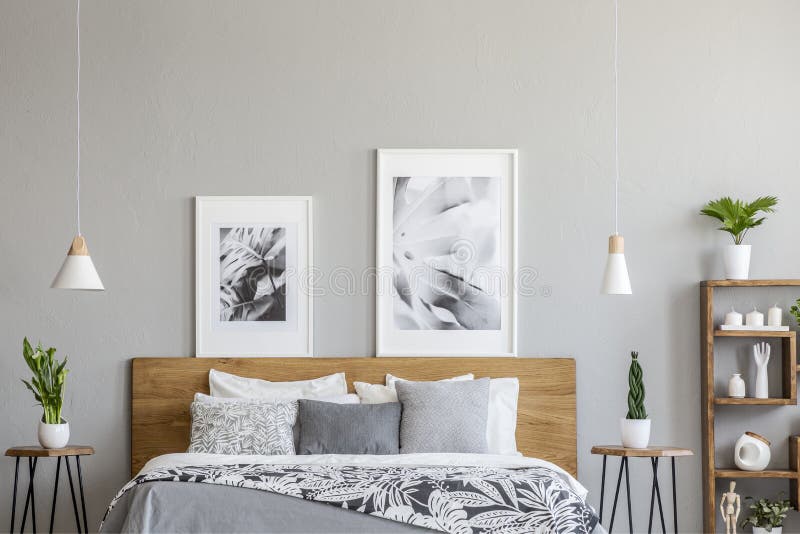 Plakaty nad drewniany łóżko między stołami z roślinami w popielatym sypialni wnętrzu z lampami Istna fotografia obraz stock Plakaty nad drewniany łóżko między stołami z roślinami w popielatym sypialni wnętrzu z lampami Istna fotografia obraz stock