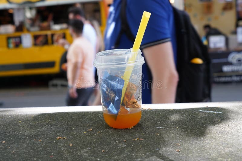 Zurich, Switzerland, 07 02 2023, Plastic glass with orange drink, straw and small pieces of wrappers from chocolate lying on table as garbage. On the background there is street with defocused incidental people passing by. Zurich, Switzerland, 07 02 2023, Plastic glass with orange drink, straw and small pieces of wrappers from chocolate lying on table as garbage. On the background there is street with defocused incidental people passing by.