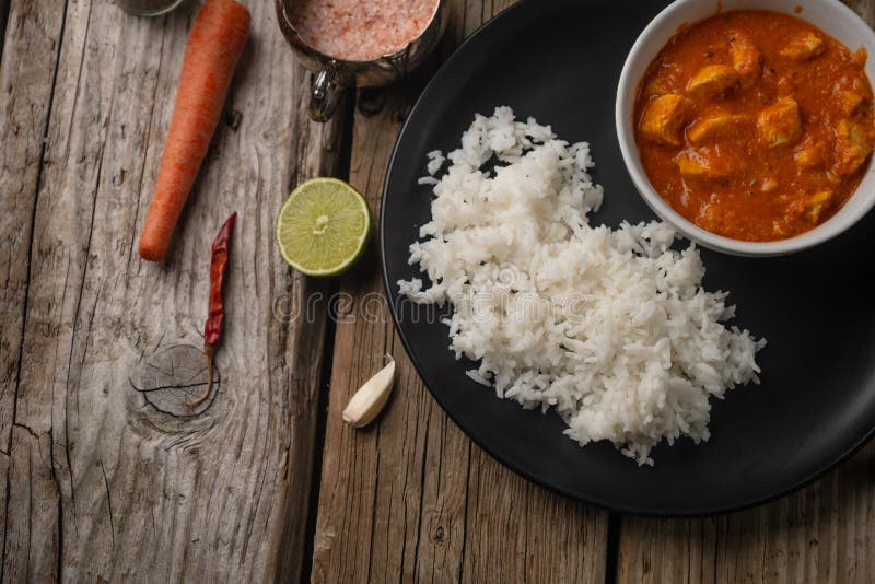 Plate with classic chicken curry and rice served with lime, garlic and red hot chilli, carrot, onion and spices on rustic wooden table background. Traditional indian cuisine. Restaurant recipe.