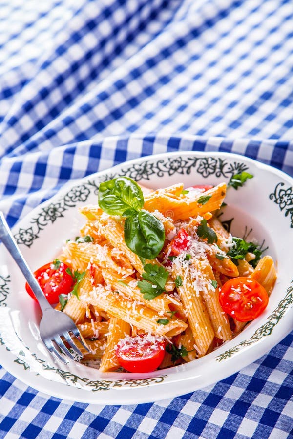 Plate with pasta pene Bolognese sauce cherry tomatoes parsley top and basil leaves on checkered blue tablecloth.