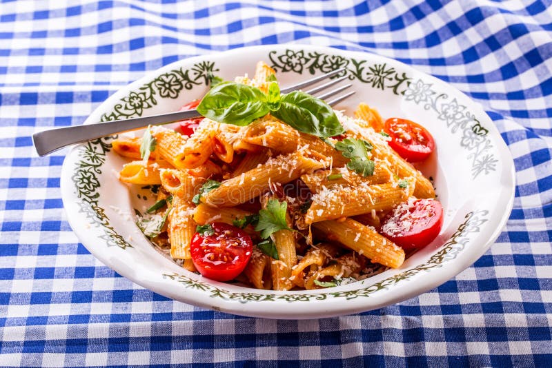 Plate with pasta pene Bolognese sauce cherry tomatoes parsley top and basil leaves on checkered blue tablecloth.