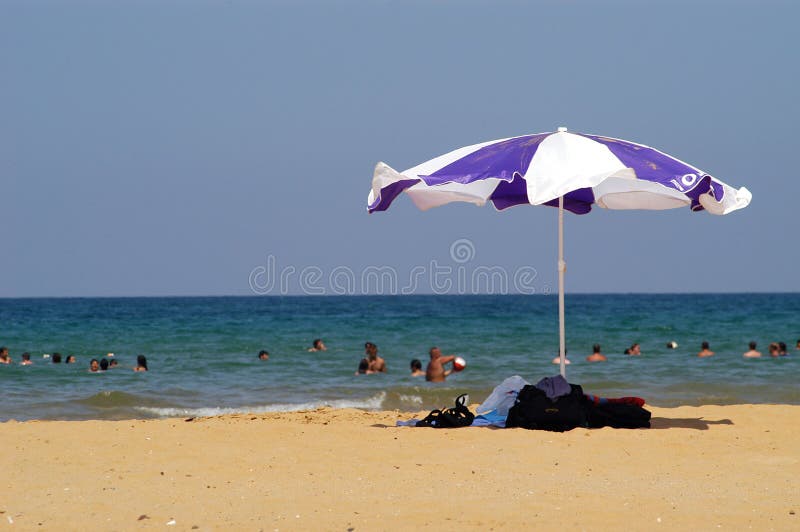 A view of a group of people swimming at a beach in Turkey on a hot, sunny day with a large white and purple beach umbrella in the foreground. A view of a group of people swimming at a beach in Turkey on a hot, sunny day with a large white and purple beach umbrella in the foreground.