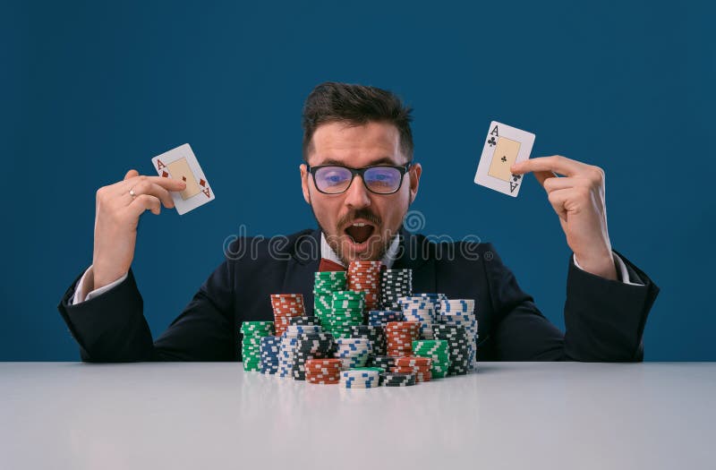 Player in glasses, black suit sitting at table with stacks of chips, holding two playing cards, posing on blue stock photo