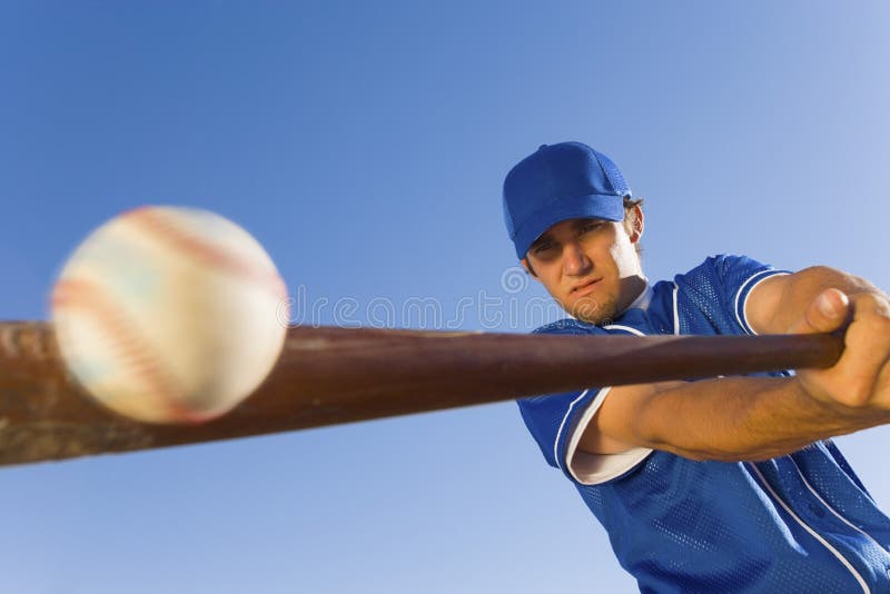 Baseballový hráč udeří míč s pálkou proti jasně modré obloze.