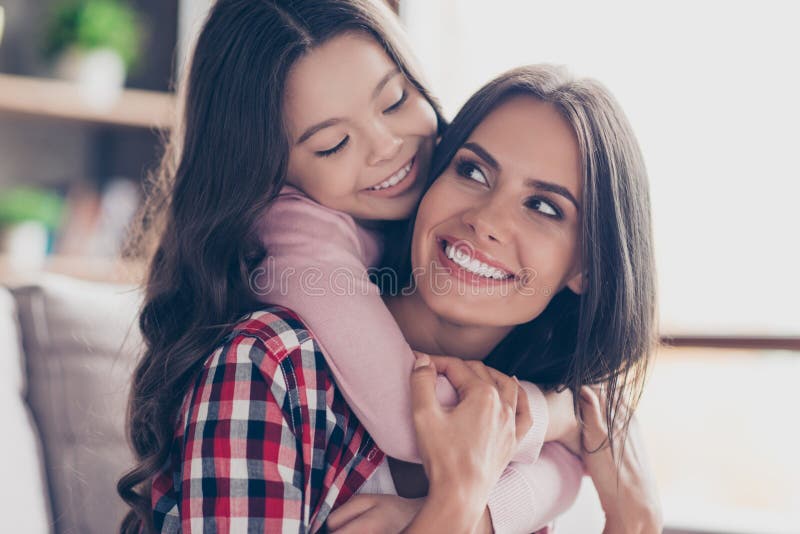 Playful small girl with long dark hair is hugging her mum`s neck