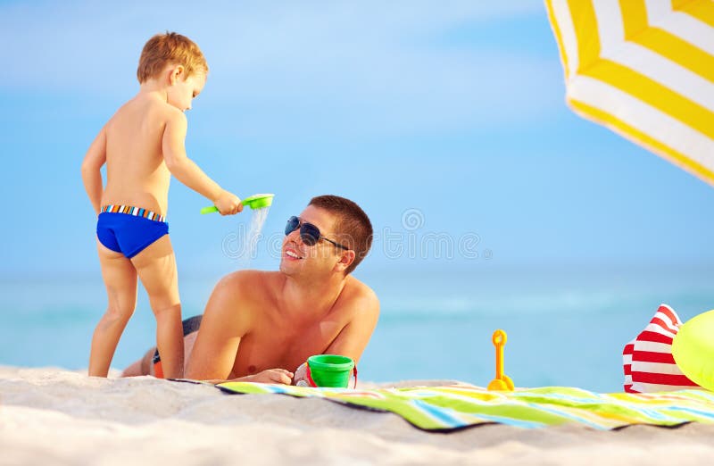 Playful son strews sand on father, colorful beach. Playful son strews sand on father, colorful beach