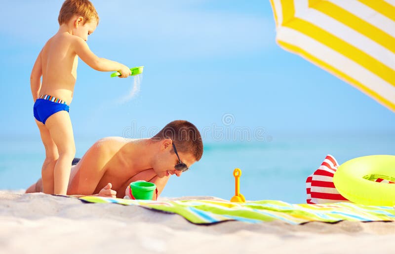 Playful son strews sand on father, colorful beach. Playful son strews sand on father, colorful beach