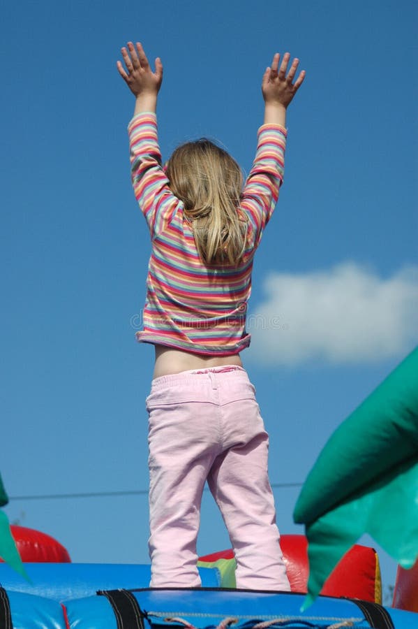 Rear view of a white caucasian girl child with long blond hair raising up her little hands in the air having great fun at playtime by standing on a jumping castle on the playground outdoors in sunshine