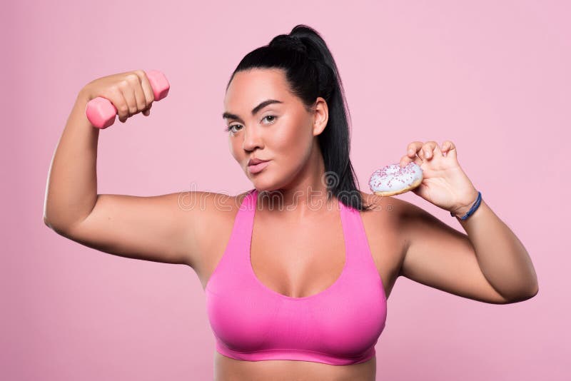Make your choice. Nice chubby woman holding dumbbell and tasty donut while standing against pink isolated background. Make your choice. Nice chubby woman holding dumbbell and tasty donut while standing against pink isolated background.