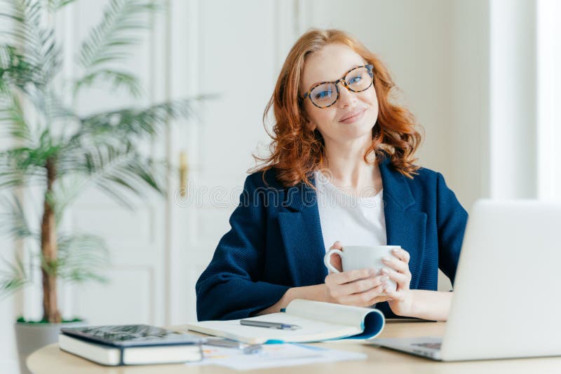 Pleased female teacher makes research work on laptop computer, writes down information in notepad, enjoys drinking coffee, has