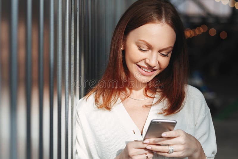 Pleased woman focused into screen of cell phone, checks email box, dressed in white clothes, sends feedback, connected to wireless