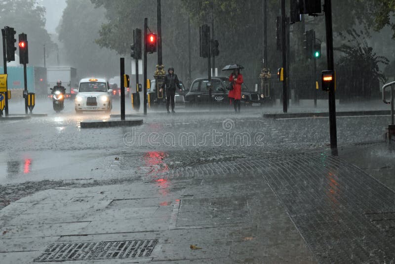 Embankment, London. Women with umbrella and anorak waiting at traffic lights, London taxi and other traffic. Dark, torrential rain. Flash flood concept, illustration. Embankment, London. Women with umbrella and anorak waiting at traffic lights, London taxi and other traffic. Dark, torrential rain. Flash flood concept, illustration.