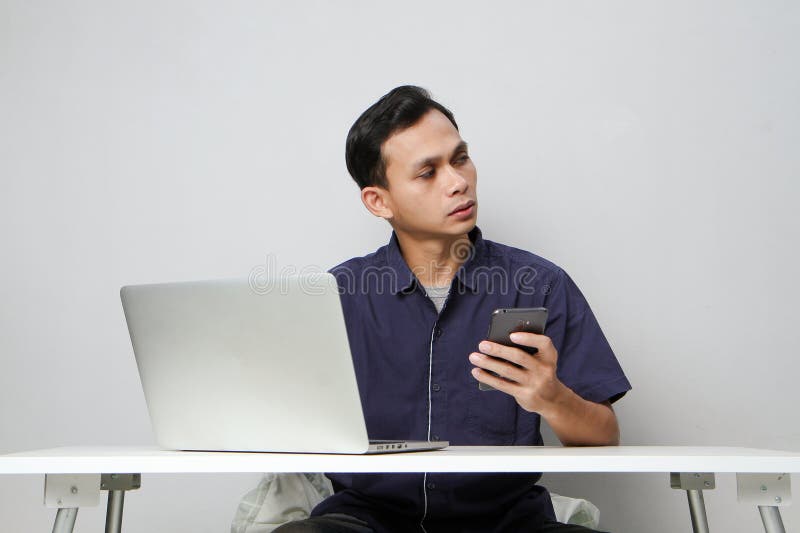 Plötslig, förvirrad som man som håller mobiltelefon medan man sitter framför den bärbara datorn. isolerad bakgrund royaltyfria bilder