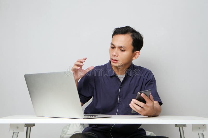 Plötslig, förvirrad som man som håller mobiltelefon medan man sitter framför den bärbara datorn. isolerad bakgrund arkivbild