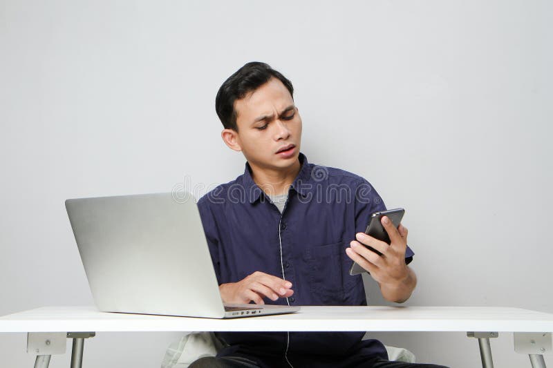 Plötslig, förvirrad som man som håller mobiltelefon medan man sitter framför den bärbara datorn. isolerad bakgrund royaltyfri bild