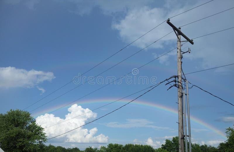 Po Podeszczowego/Podwaja tęczę, zwierzęcia, linii energetycznych/. Niebieskie niebo nad treetops z dwoistą tęczą arced przez; Puszyści biel chmury spojrzenia jak obsiadanie pies, lew lub inny zwierzęcy strzeżenie garnek złoto przy tęczy końcówką; niebo elektryfikuje wysokimi drutami