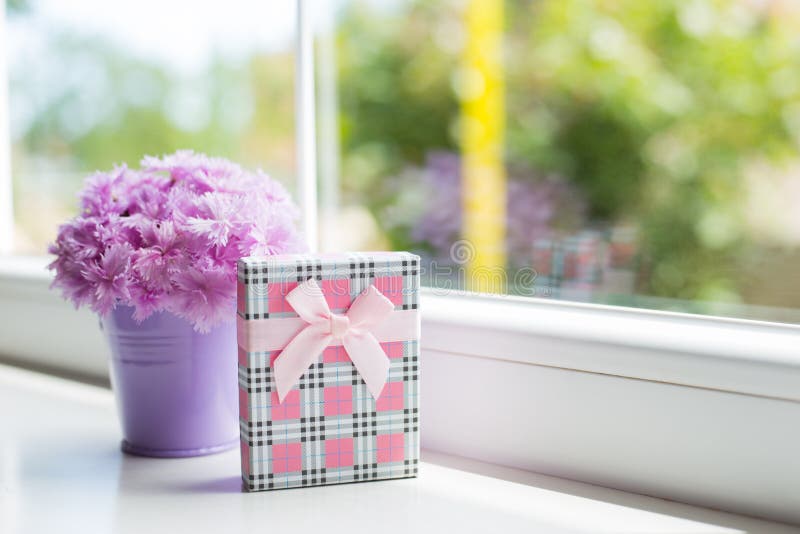 Little purple bucket with tender bouquet of beautiful pink carnation with pink gift box near window in daylight. Spring card. Little purple bucket with tender bouquet of beautiful pink carnation with pink gift box near window in daylight. Spring card.