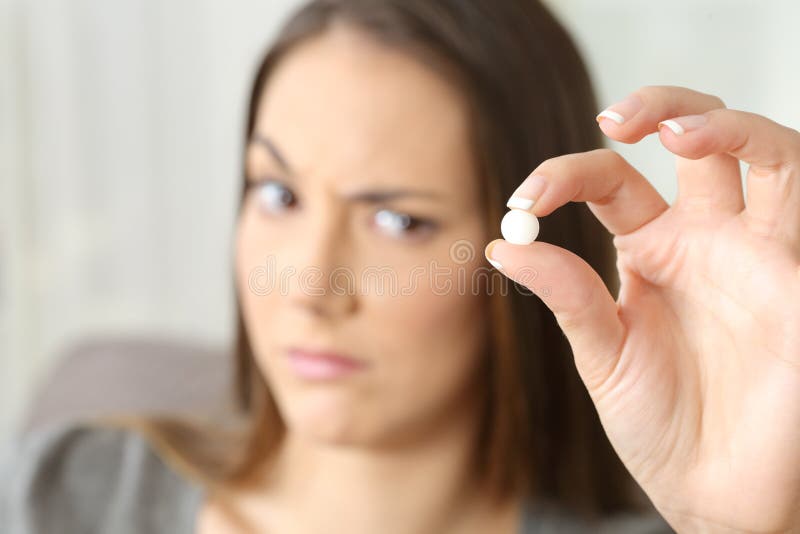 Suspicious woman looking at a white round pill sitting on a sofa in a house interior. Suspicious woman looking at a white round pill sitting on a sofa in a house interior