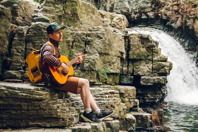 Podróżnika mężczyzna z plecakiem bawić się gitarę przeciw siklawie Przestrze? dla tw?j promocyjnej zawarto?ci lub wiadomo?ci teks zdjęcie stock Podróżnika mężczyzna z plecakiem bawić się gitarę przeciw siklawie Przestrze? dla tw?j promocyjnej zawarto?ci lub wiadomo?ci teks zdjęcie stock